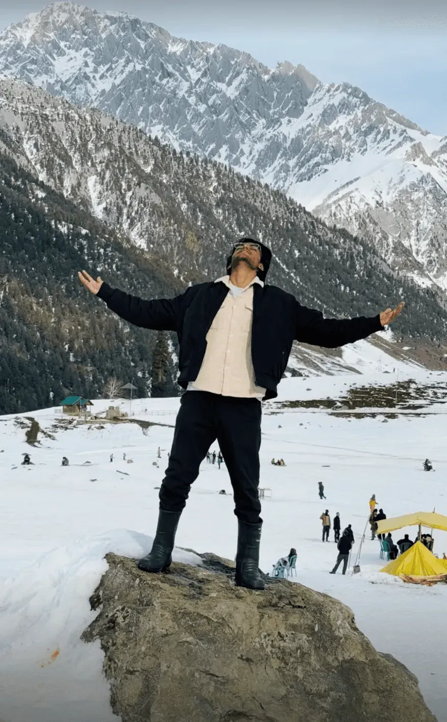 Standing on a boulder with both arms out, face to the sky, snowfield and peaks behind