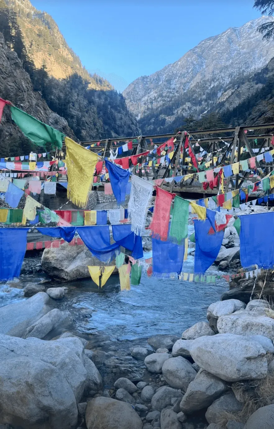 Rows of prayer flags strung across a wooden footbridge over a boulder-strewn river
