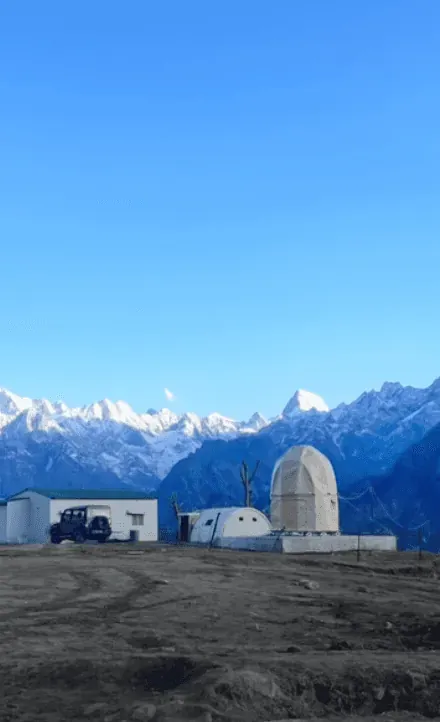A small camp of huts on a bare ridge with a snow-covered range behind it
